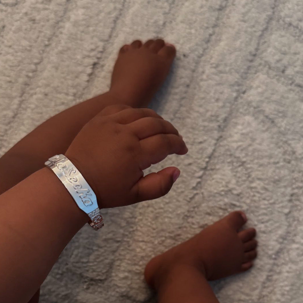 Baby wearing a personalized sterling silver name bracelet on the wrist, photographed on a soft neutral textile background.