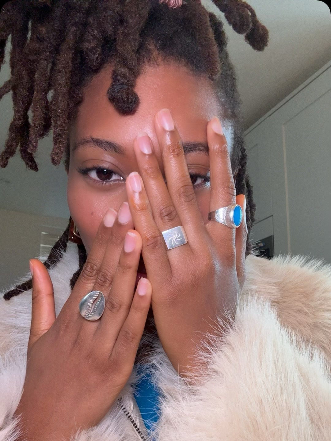 Person with styled hair wearing rings, covering face with hands indoors.