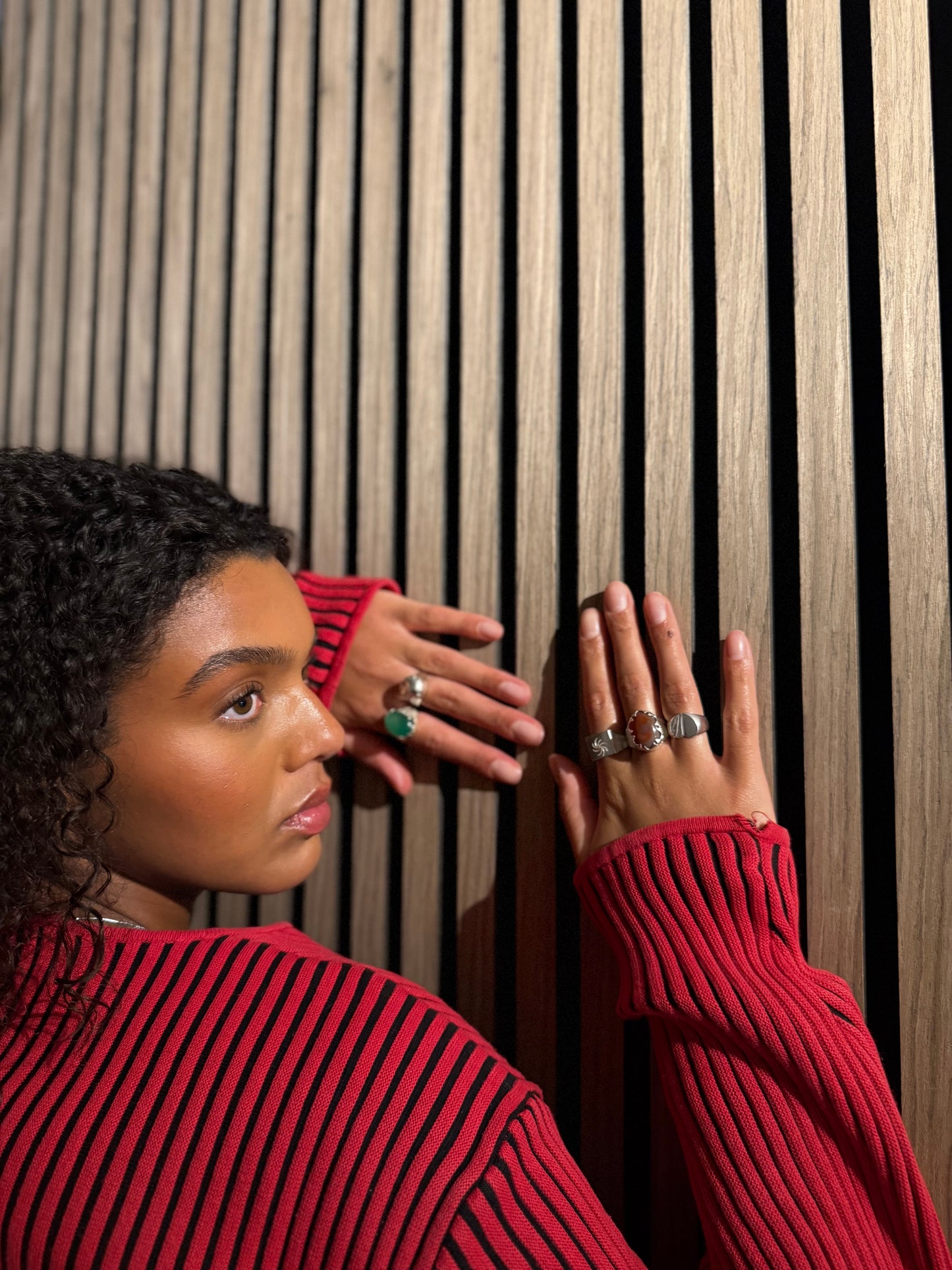 Woman wearing a red sweater with multiple rings, looking away from the camera against a wooden slat background.
