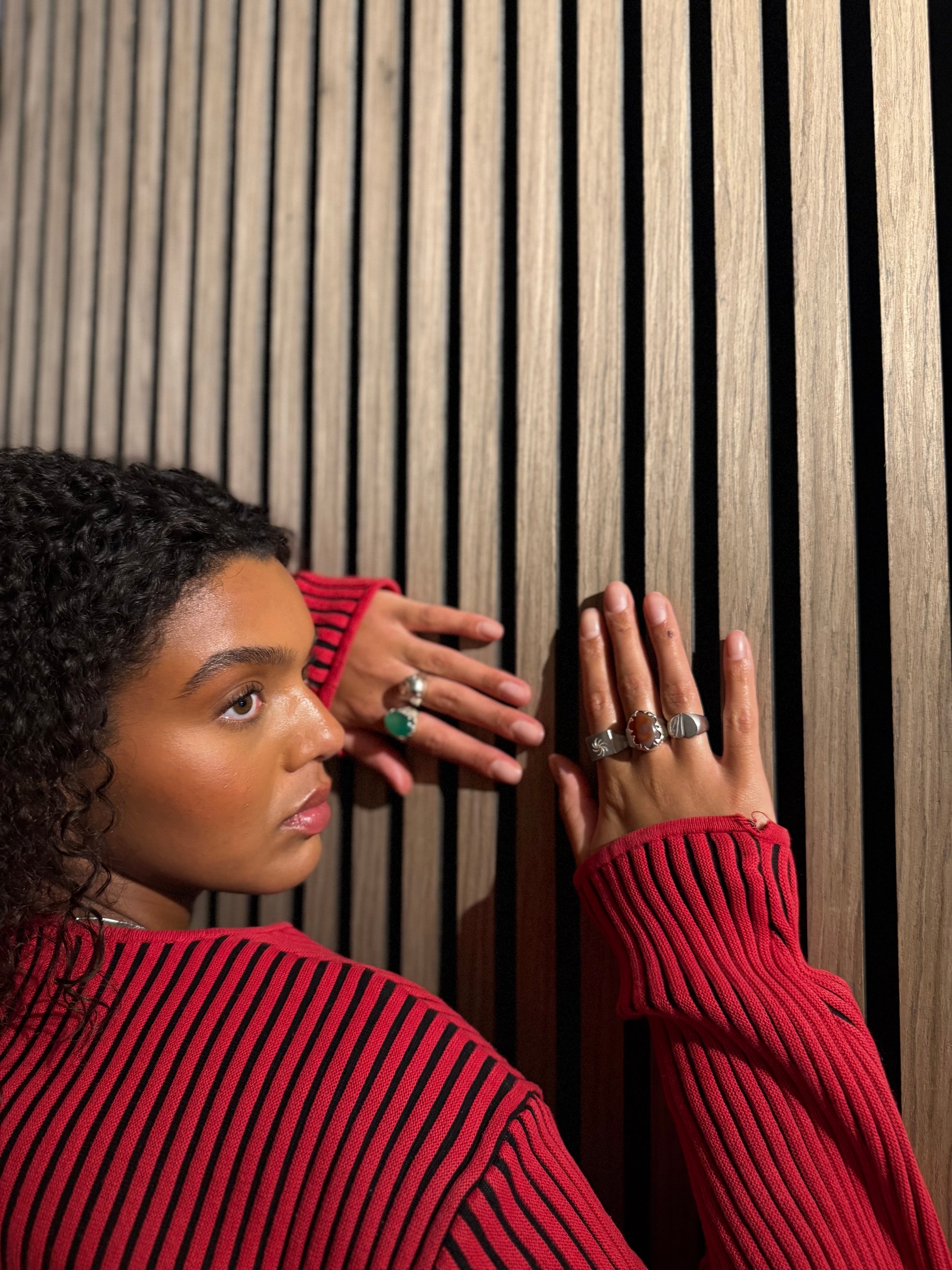 Woman wearing a red sweater with multiple rings, looking away from the camera against a wooden slat background.